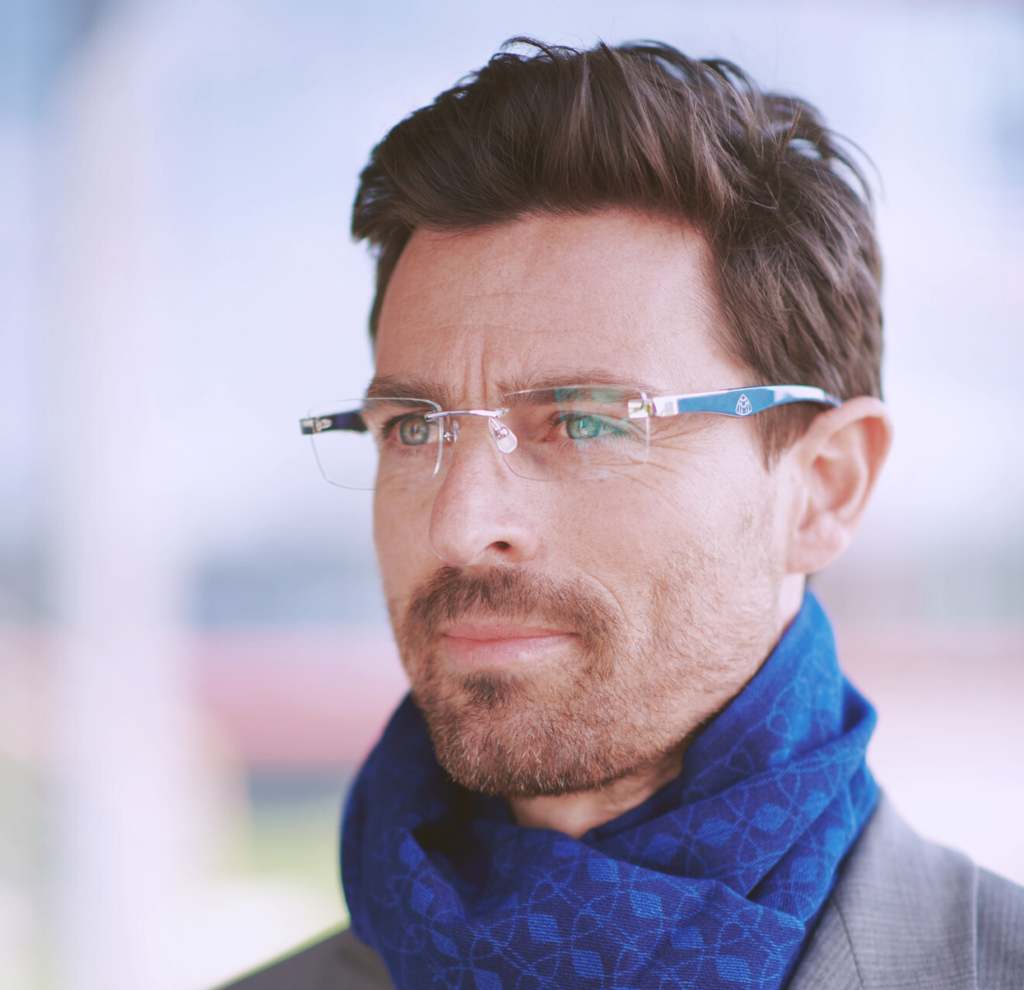 A distinguished man with neatly styled hair and a trimmed beard is depicted gazing thoughtfully into the distance. He is wearing rimless, rectangular R&B Maybach eyeglasses that feature a minimalist design with subtle detailing on the temples. A luxurious blue scarf with an intricate geometric pattern is wrapped around his neck, complementing the elegant and sophisticated aura that the glasses add to his smart, grey suit. The softly focused background suggests an urban setting, highlighting the glasses' suitability for a modern, professional lifestyle.