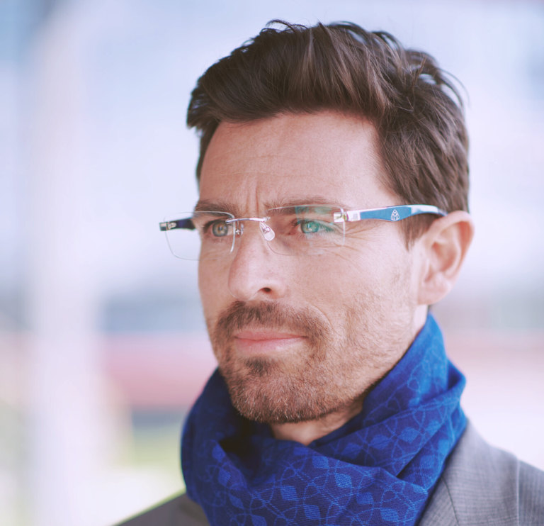 A distinguished man with neatly styled hair and a trimmed beard is depicted gazing thoughtfully into the distance. He is wearing rimless, rectangular R&B Maybach eyeglasses that feature a minimalist design with subtle detailing on the temples. A luxurious blue scarf with an intricate geometric pattern is wrapped around his neck, complementing the elegant and sophisticated aura that the glasses add to his smart, grey suit. The softly focused background suggests an urban setting, highlighting the glasses' suitability for a modern, professional lifestyle.