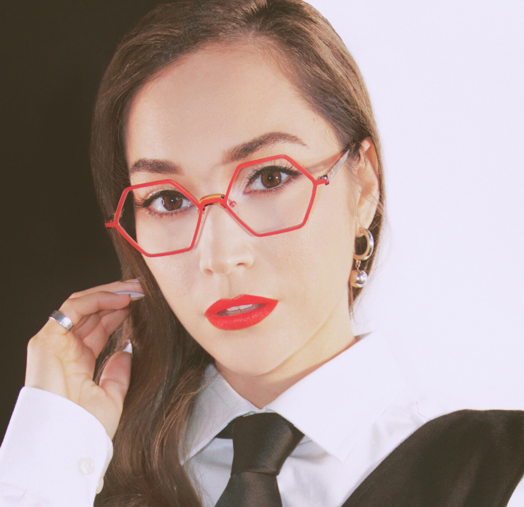 A confident woman with dark hair is portrayed looking directly at the viewer, wearing distinctive red, hexagon-shaped I A Eyeworks glasses. The frames make a bold statement against her fair complexion, complemented by her striking red lipstick. She is dressed in a classic white shirt with a black tie, which along with her subtle earrings and ring, adds a touch of elegance to her professional appearance. The high-contrast lighting accentuates her features and the vivid color of the eyewear, highlighting the glasses as a fashionable accessory for a stylish and assertive look.