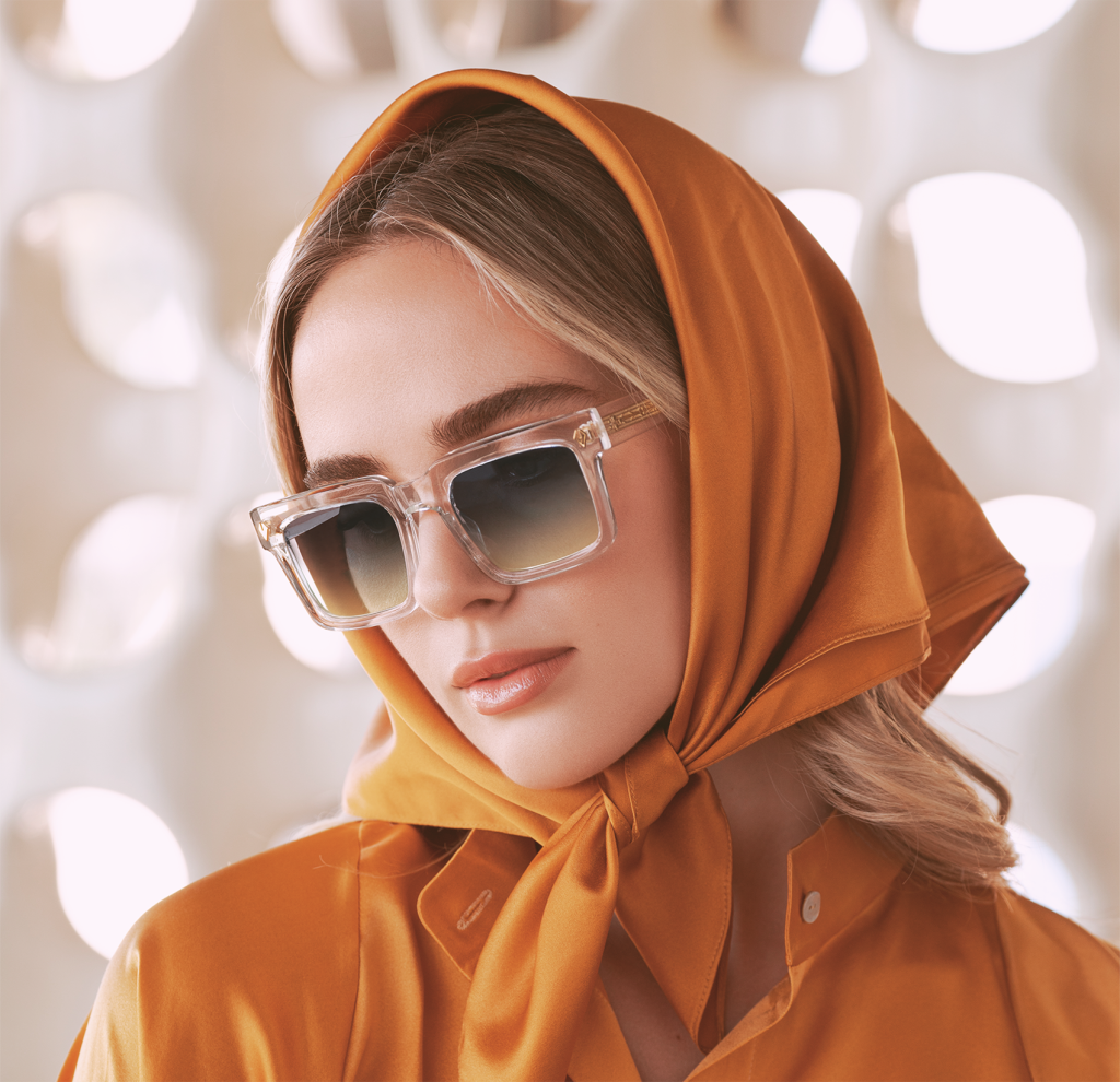 A woman with an orange scarf and a burnt orange shirt is wearing a pair of large, square-framed, clear T Henri sunglasses with gradient lenses. The glasses have a bold, modern design with gold accents on the hinges, showcasing a luxurious and contemporary style. The background is softly blurred with geometric patterns that complement her chic and sophisticated look.