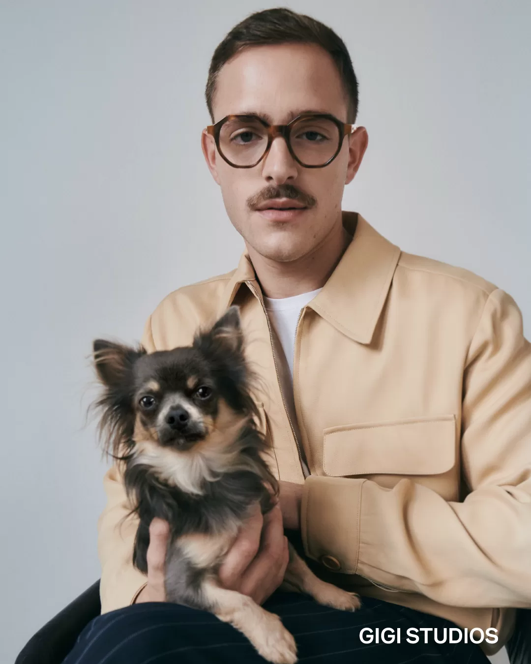 A fashionable man with a neat mustache and wearing stylish, large square-framed glasses poses with a small Chihuahua in his arms. He is dressed in a beige jacket over a white shirt, exuding a sophisticated yet approachable vibe. The dog looks directly at the camera, adding a cute and endearing element to the image. The background is simple and light-colored, focusing attention on the subjects.