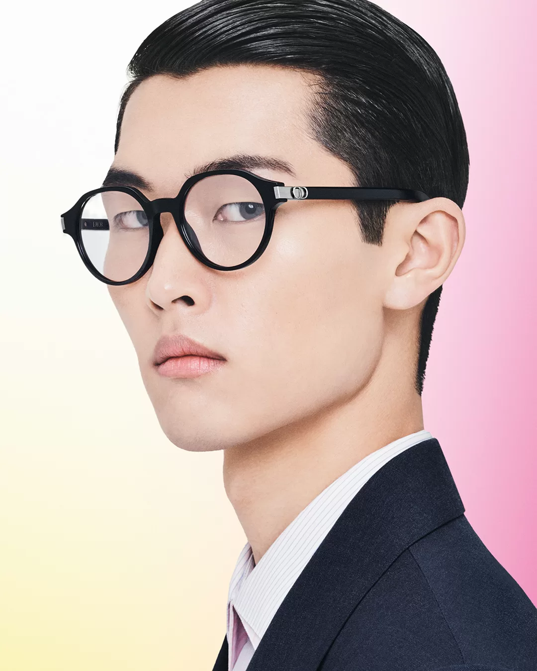 A portrait of a young Asian man in elegant attire, featuring black, thick-framed glasses. His hair is slicked back, and he wears a dark navy suit with a light blue shirt and dark tie. The background is softly divided into pastel pink and yellow.