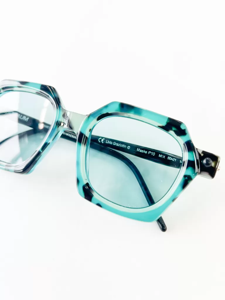 This image displays a pair of avant-garde eyeglasses lying flat against a white background. The frames are thick and made from translucent turquoise acetate, giving them a vibrant and fresh look. The shape of the glasses is a unique octagonal, merging contemporary style with a hint of retro flair. The large, clear lenses suggest the frames are designed for fashion with a practical purpose, possibly offering the wearer a broad field of vision. These eyeglasses are modern, yet timeless, appealing to those who favor bold, statement accessories.