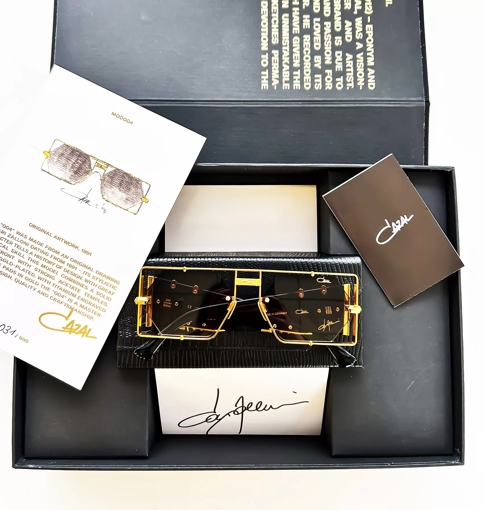 A pair of Cazal sunglasses displayed in a black presentation box, accompanied by a designer card, a sketch of the glasses, and a description booklet.