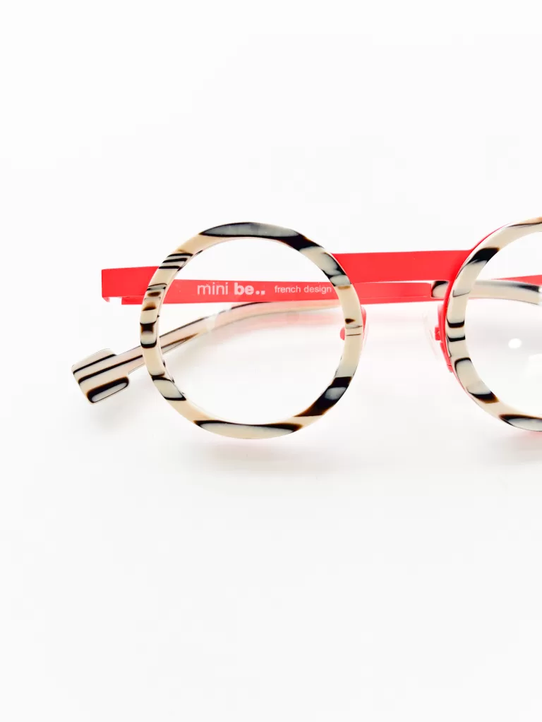This image features a pair of stylish children's eyeglasses with a unique round frame design. The glasses have a playful tortoiseshell pattern with splashes of black, beige, and ivory. A striking red band runs across the top of the frame, emblazoned with the words "mini be.. French design," indicating the brand and its origin. The eyeglasses are positioned flat against a pure white background, emphasizing their colorful and vibrant design, ideal for a young wearer looking to add a touch of fun and fashion to their daily wear.