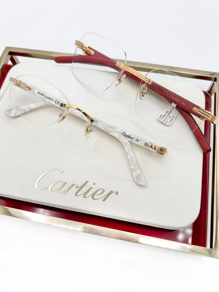 Elegant Cartier eyeglasses presented on a cream and red display box. The glasses feature gold accents and transparent frames with minimalistic, rimless design, emphasizing their sophisticated craftsmanship.