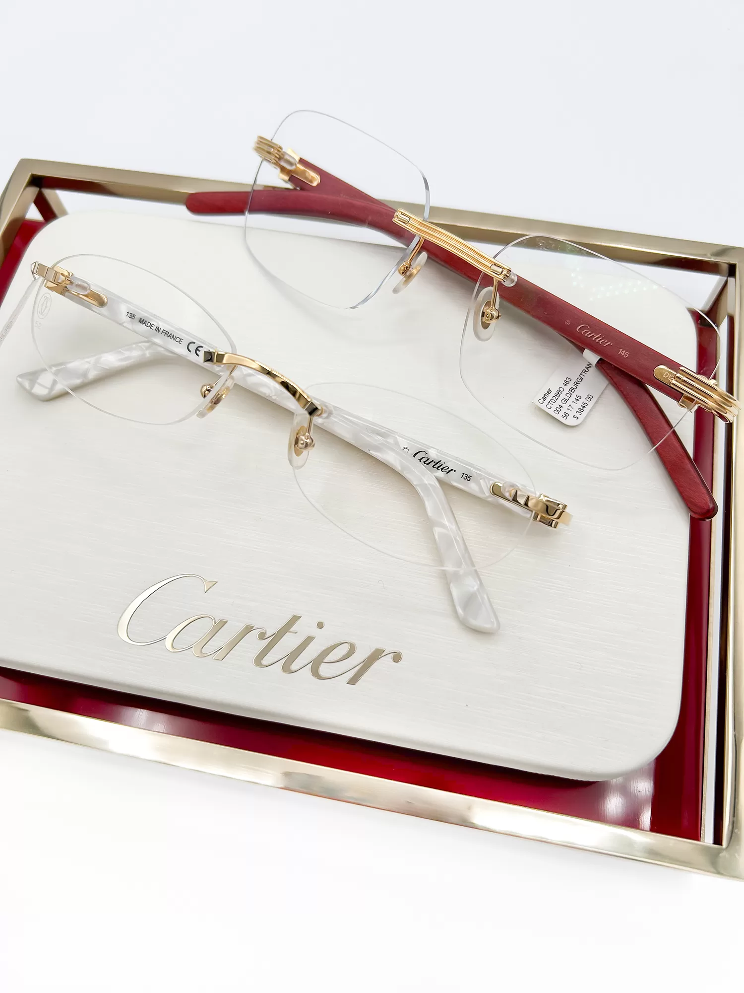 Elegant Cartier eyeglasses presented on a cream and red display box. The glasses feature gold accents and transparent frames with minimalistic, rimless design, emphasizing their sophisticated craftsmanship.