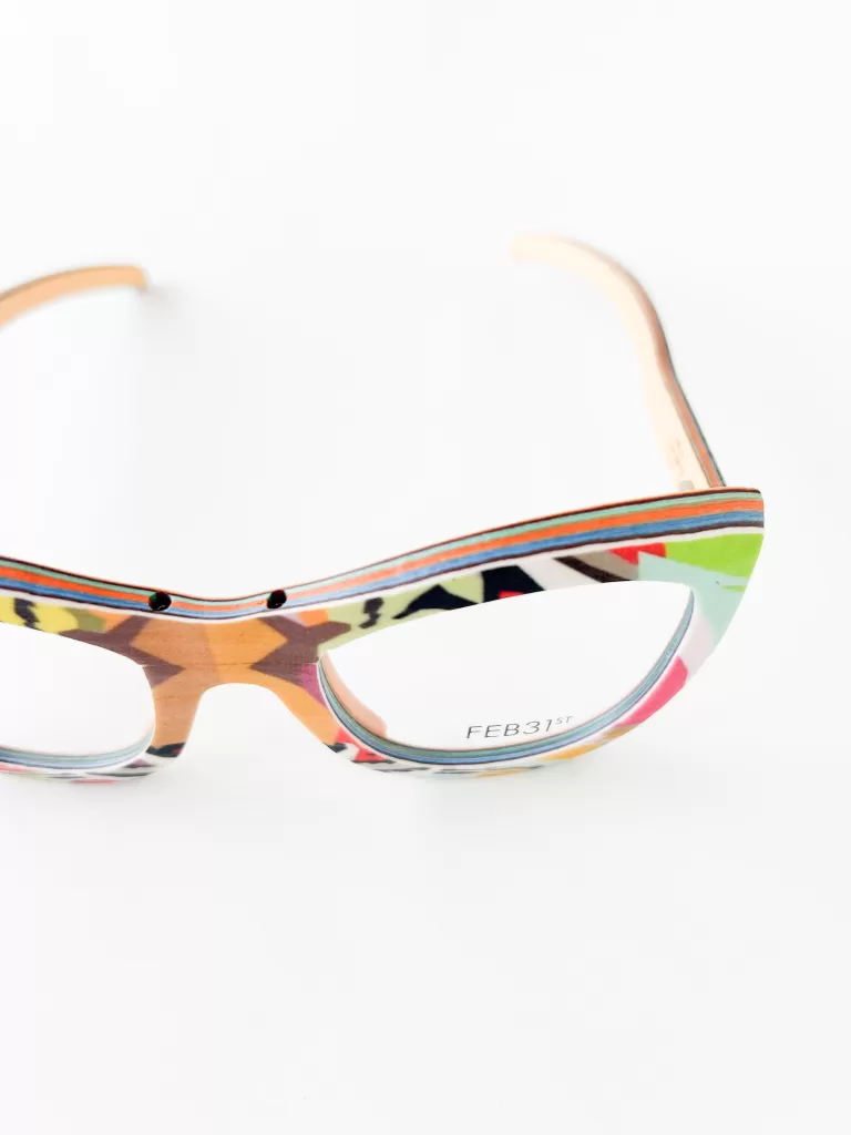This image displays a pair of eyeglasses with a uniquely artistic and colorful frame. The frame features a vibrant and eclectic mix of patterns, incorporating various hues such as red, blue, green, and yellow, set against a beige base. This distinctive pattern makes the glasses not only a visual accessory but also a statement piece. The temples of the glasses have a natural wood finish, adding an element of organic texture and contrast to the colorful frame. The design is marked with the branding "FEB31st," indicating a focus on craftsmanship and originality. Set against a white background, the glasses are positioned to showcase their dynamic and unconventional style, appealing to those who appreciate eyewear as an expression of individuality and art.