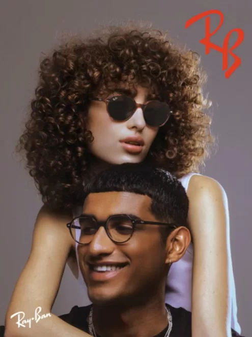 A stylized fashion portrait featuring two individuals showcasing Ray-Ban eyewear. The woman, with voluminous curly hair, sports classic round, metal-framed sunglasses and rests atop the shoulder of a smiling man wearing sleek, rectangular eyeglasses. Both exhibit a cool, relaxed demeanor against a soft gray background.