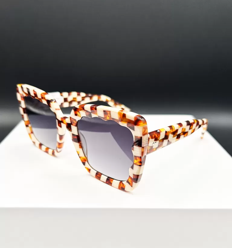 Elegant tortoiseshell sunglasses with a distinct and angular frame design, showcased on a white pedestal against a dark background. The glasses feature thick frames with a complex pattern of amber, brown, and honey hues, enhancing their stylish appearance.