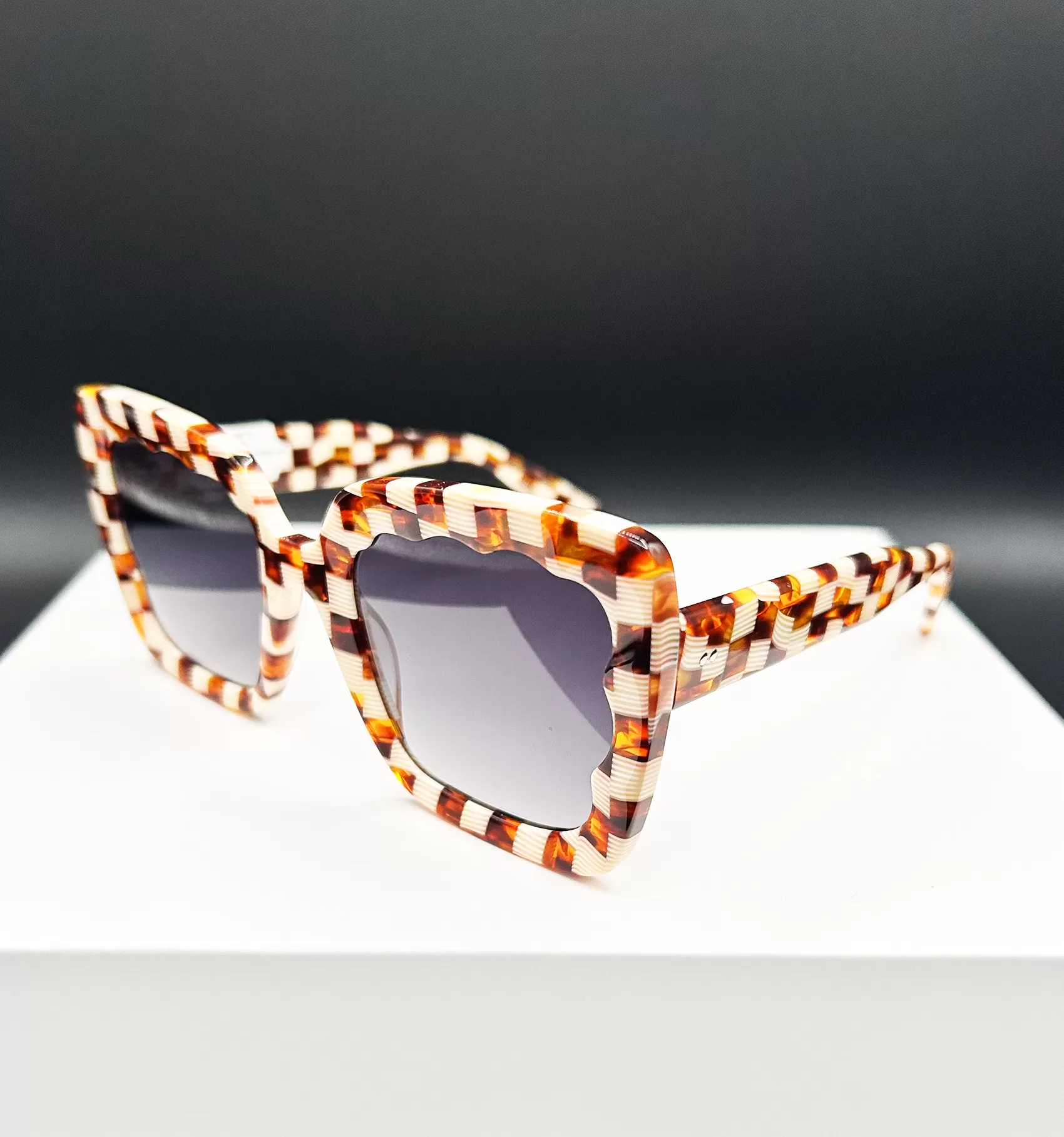 Elegant tortoiseshell sunglasses with a distinct and angular frame design, showcased on a white pedestal against a dark background. The glasses feature thick frames with a complex pattern of amber, brown, and honey hues, enhancing their stylish appearance.