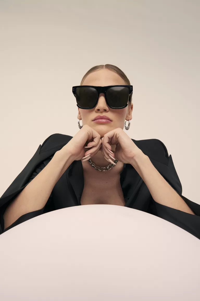 A fashion portrait of a woman wearing oversized black sunglasses with a bold frame. She is dressed in a sophisticated black outfit with a prominent black blazer, and accessorized with large hoop earrings and a chunky silver necklace. She rests her chin on her hands atop a pink circular table, against a soft beige background.