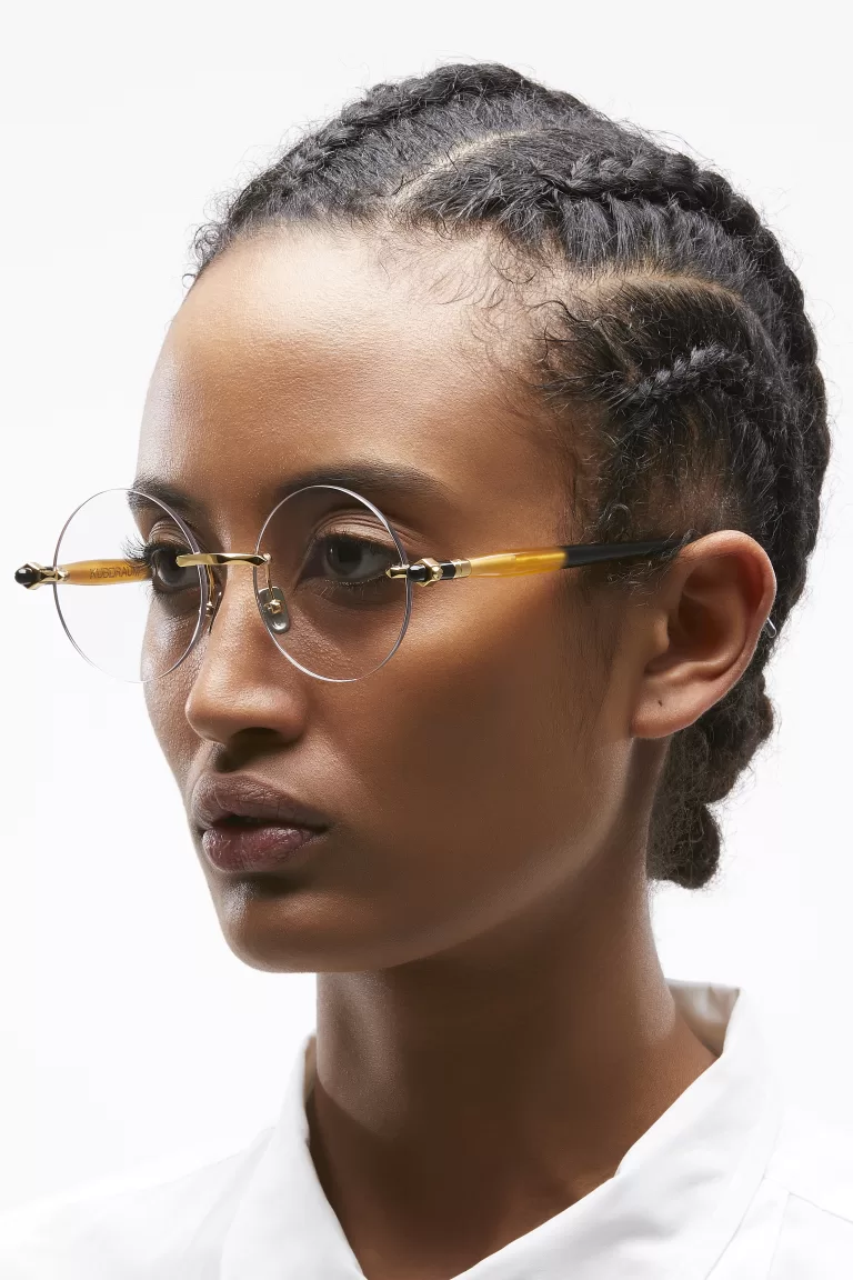 Portrait of a young woman with intricate braided hair, wearing delicate round-frame gold and caramel glasses by Kuboraum. The glasses are minimal yet sophisticated, emphasizing her confident gaze.