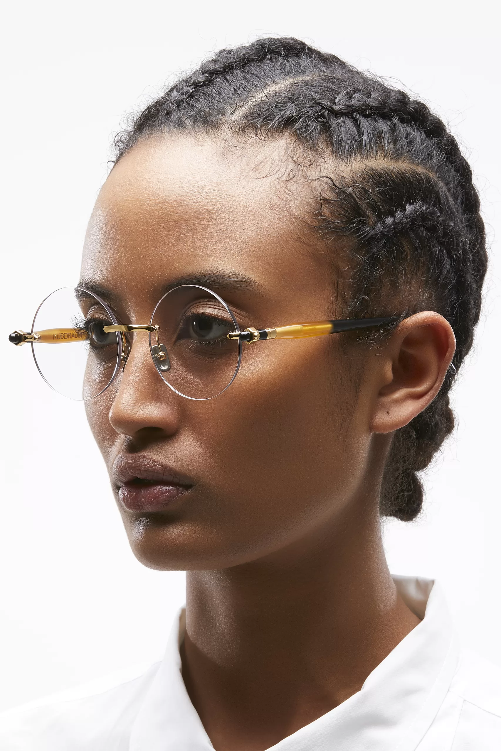 Portrait of a young woman with intricate braided hair, wearing delicate round-frame gold and caramel glasses by Kuboraum. The glasses are minimal yet sophisticated, emphasizing her confident gaze.