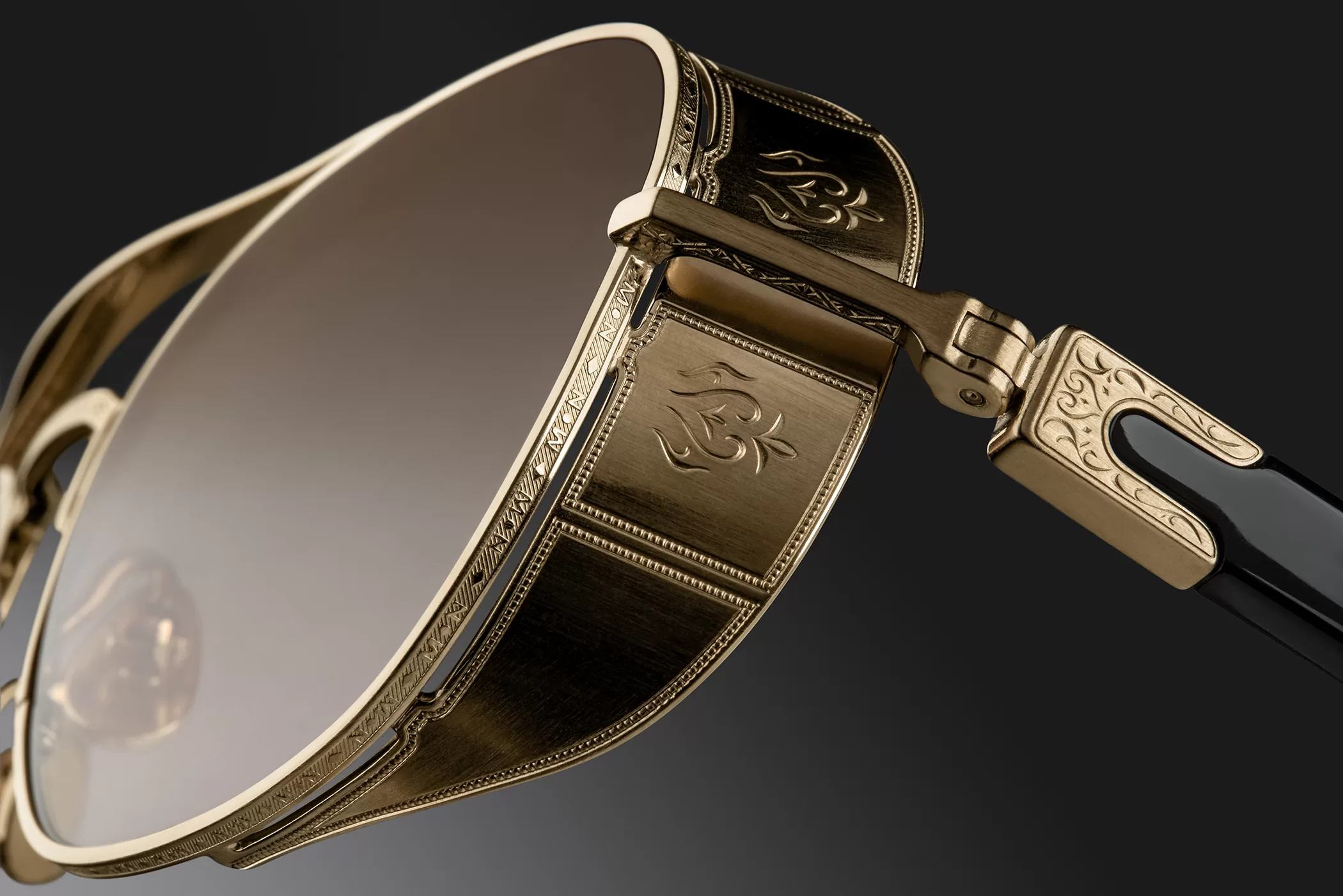 This image features a close-up of a luxurious pair of Matsuda sunglasses. The frame is crafted from polished gold metal with intricate engraving details throughout, showcasing traditional Japanese artistry. The hinges are adorned with a decorative floral motif that adds an elegant touch. The lenses are large and oval with a subtle gradient tint, blending seamlessly into the ornate frame. The arms of the glasses are also gold with black acetate tips for comfort. This pair of sunglasses combines functionality with high-end craftsmanship, reflecting a sophisticated and refined style.
