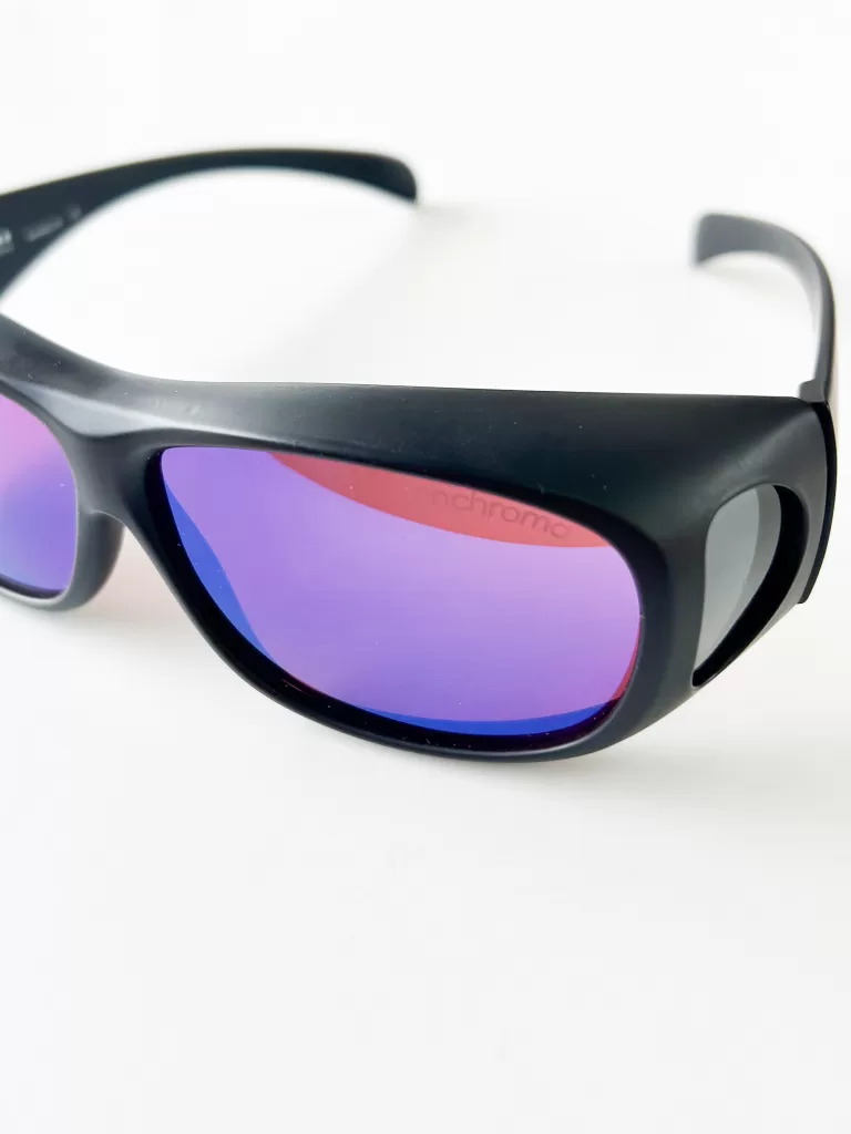 This image showcases a pair of specialized eyeglasses designed for color vision correction, prominently featuring a matte black frame with ergonomic design for comfortable wear. The lenses are notable for their striking violet tint, which aids in enhancing color perception for colorblind individuals. The sleek, sporty style of the frame, combined with the advanced lens technology, makes these glasses not only a functional tool for those with color vision deficiencies but also a stylish accessory. The background is pure white, emphasizing the vibrant color and clarity of the lenses.