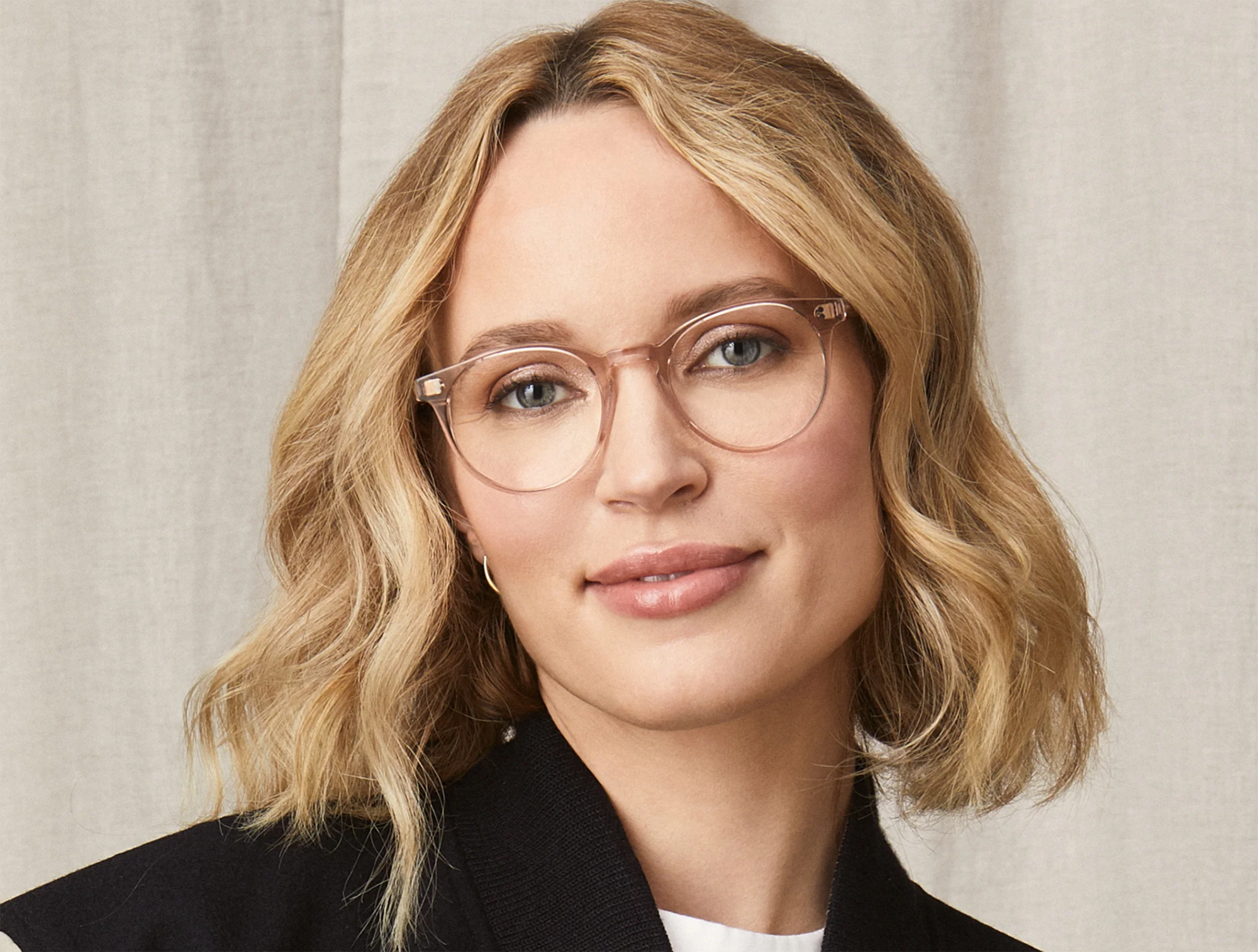 Portrait of a woman with shoulder-length wavy blonde hair, wearing clear round glasses with a thin metallic frame. She has a subtle smile and is dressed in a black blazer over a white blouse, set against a beige textured background.