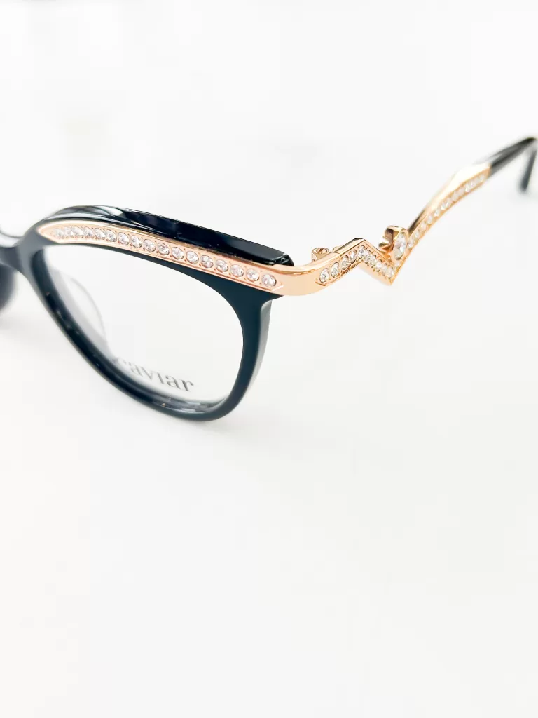 This image showcases an exquisite pair of eyeglasses that blend opulence with functionality. The frame is predominantly black with a classic oval shape, crafted from high-quality acetate for a sleek and polished look. The standout feature of these glasses is the luxurious gold embellishment along the temples, intricately detailed and studded with a row of sparkling crystals. This embellishment adds a glamorous touch, making the glasses not just a vision accessory but a statement piece of jewelry. The contrasting materials and the blend of simplicity with luxury make these glasses ideal for those who appreciate eyewear that serves as a focal point of sophistication and style. The white background enhances the vividness of the golden accents and the clarity of the crystals, highlighting their meticulous craftsmanship.