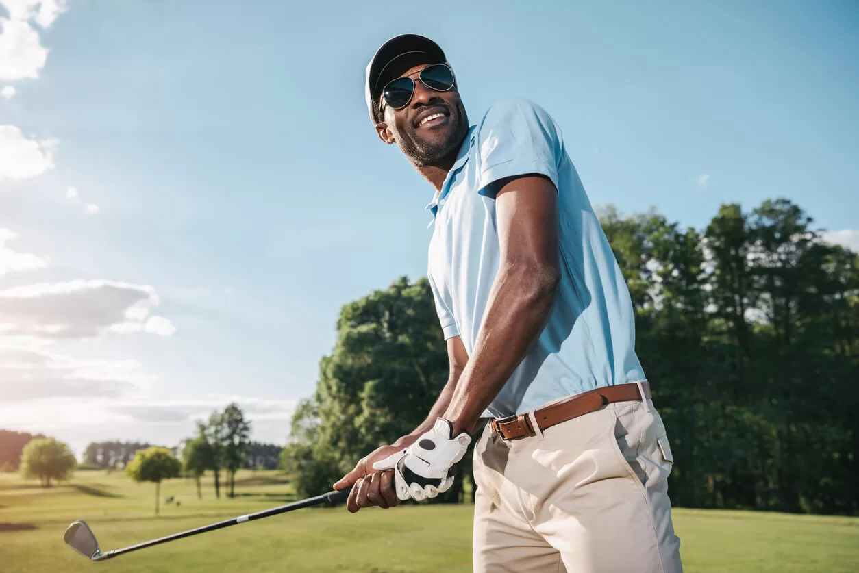 Smiling African American man in cap and sunglasses holding club and playing golf